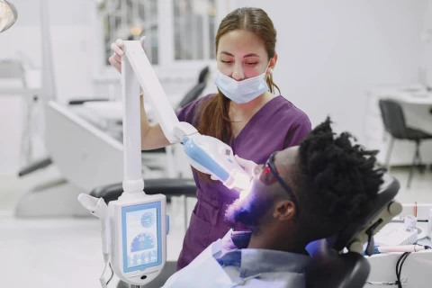 Dentist performing a teeth whitening procedure on a patient wearing protective glasses, using a specialized light device in a modern dental clinic.