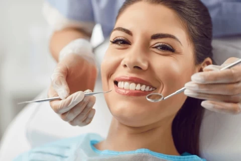 Smiling woman receiving a dental checkup, showcasing healthy teeth, with a dentist holding dental tools in the background.