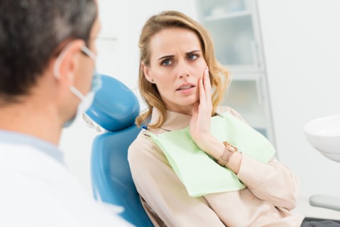 Concerned female patient holding her cheek, indicating toothache, while consulting with a dentist in a modern dental clinic.